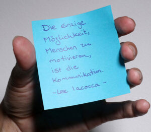 Eine Hand hält einen blauen Zettel mit einem Zitat zu Kommunikation.