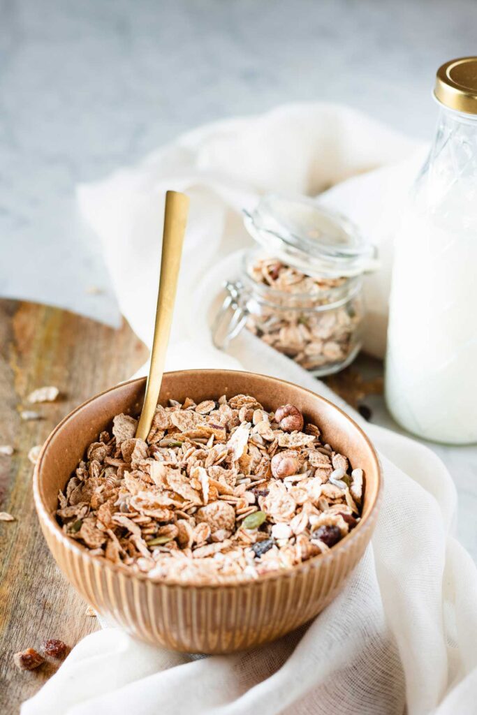 Schüssel mit Müsli und Haferflochen, goldener Löffel und Milchflasche im Hintergrund